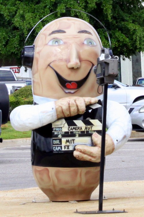 A large, cartoonish peanut character wearing headphones, holding a film clapperboard, with a cheerful expression.