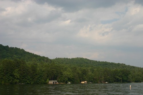 A serene lake scene with green hills, cloudy skies, and small boathouses along the water's edge.