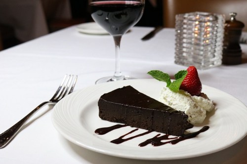 A slice of rich chocolate cake with whipped cream, a strawberry, and a glass of red wine on a white tablecloth.