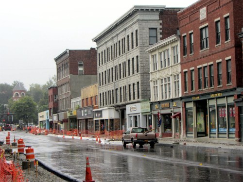 A rainy street scene with historic buildings, construction barriers, and a parked car in a small town.