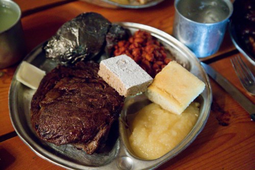 A metal plate with a large piece of meat, cornbread, applesauce, and a side of beans, set on a wooden table.
