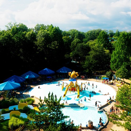 Aerial view of a water park with a circular pool, colorful umbrellas, and people enjoying the water on a sunny day.
