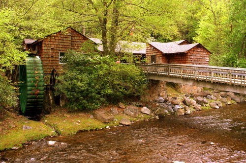 A rustic watermill beside a stream, with a wooden bridge and lush green trees surrounding the scene.