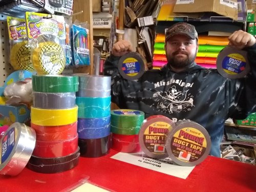 A man holds two rolls of duct tape, surrounded by various colors of tape stacked on a table in a store.