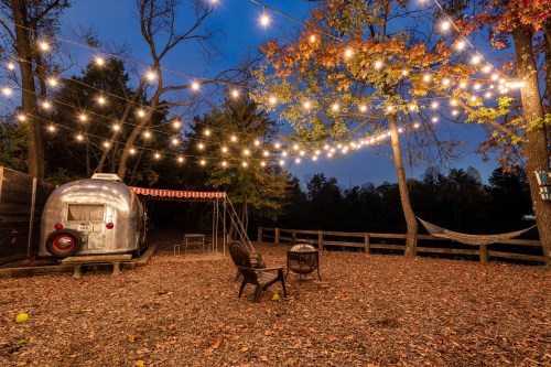 A cozy outdoor space with string lights, a vintage trailer, and seating surrounded by autumn leaves and trees.