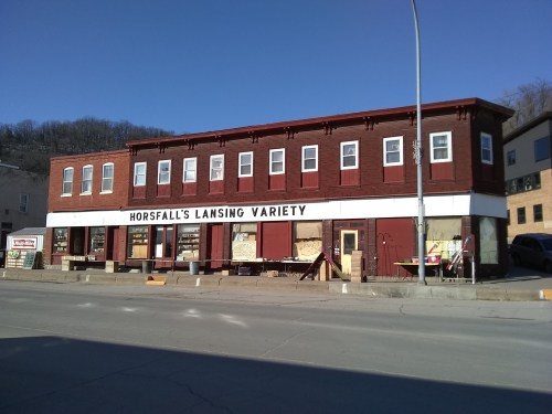 A historic two-story building with a red facade, featuring "Horsfall's Lansing Variety" signage and large windows.