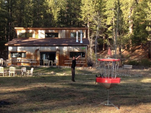 A person stands in a grassy area near a rustic building, with a disc golf basket in the foreground and trees in the background.