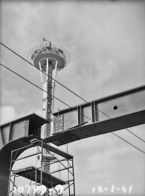 Black and white image of the Space Needle under construction, with scaffolding and steel beams in the foreground.