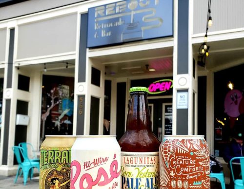 Four craft beer cans in front of a bar with a sign that reads "Reboot" and an open sign in the window.
