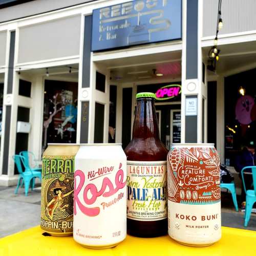 Four colorful beverage cans and a bottle are displayed outside a bar, with outdoor seating visible in the background.