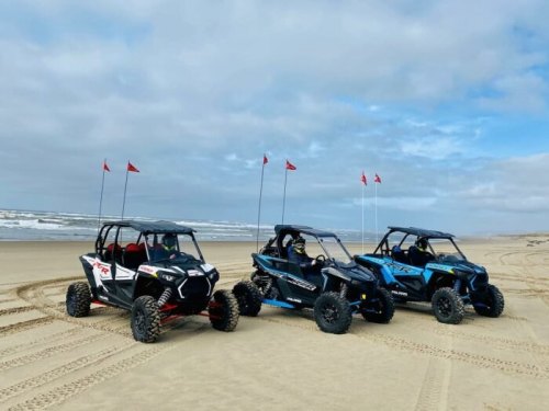Three off-road vehicles parked on a sandy beach, with ocean waves and cloudy skies in the background.