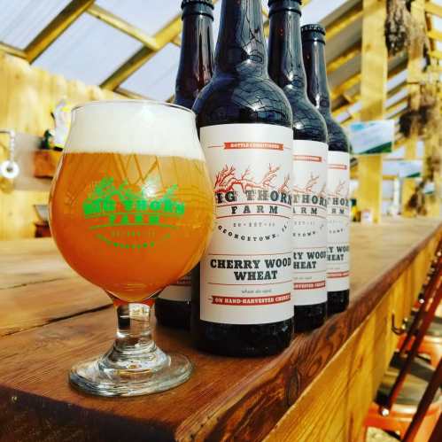 A glass of Cherry Wood Wheat beer sits on a wooden table, with bottles in the background, all in a rustic setting.