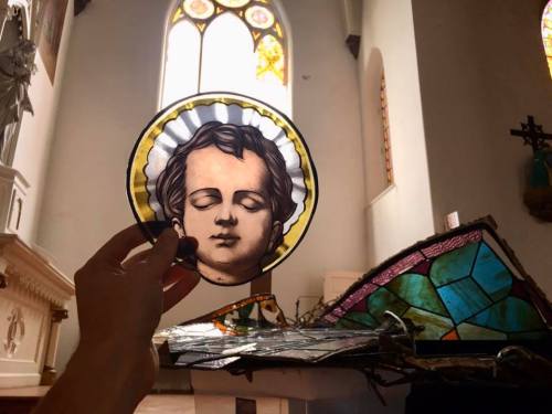 A hand holds a circular stained glass piece depicting a serene face, with a broken stained glass window in the background.