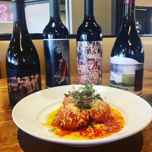 A plate of cheesy meatballs in marinara sauce, with four bottles of wine in the background.