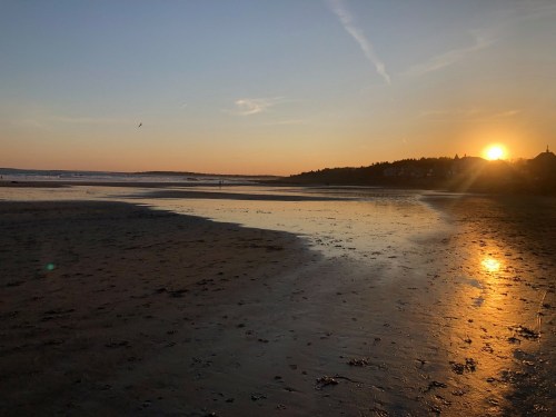 Sunset over a tranquil beach, with reflections on wet sand and a clear sky in the background.