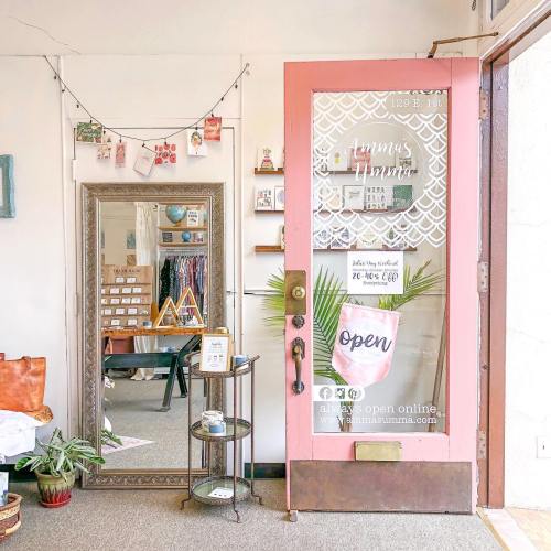 A cozy shop entrance with a pink door, mirror, and decor, featuring an "Open" sign and plants.