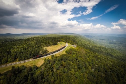Aerial view of a train traveling through lush green hills under a partly cloudy sky.