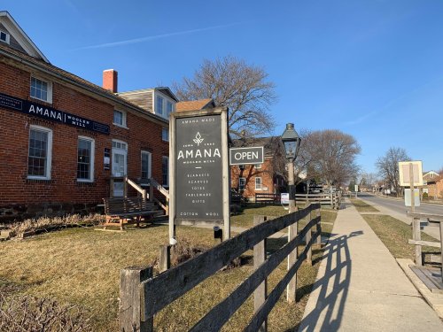 Sign for Amana Woolen Mill with "Open" displayed, surrounded by brick buildings and a clear blue sky.