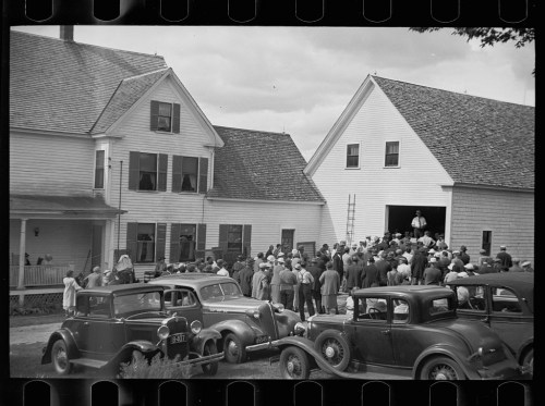 A large crowd gathers outside a house, with vintage cars parked nearby, under a cloudy sky.