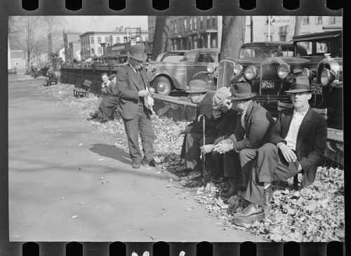 A group of men in hats sitting on a park bench, with cars and trees in the background, during autumn.