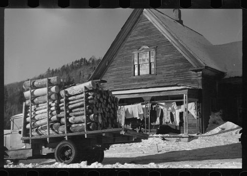 A truck loaded with logs parked in front of a wooden house, with laundry hanging outside in the snow.