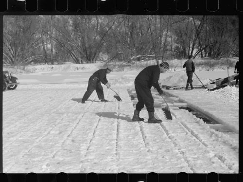 Two men shoveling snow on a frozen surface, with trees in the background and a third person working nearby.