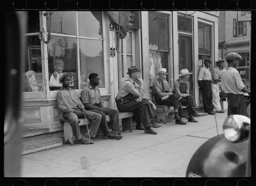 A group of men and boys sitting on a bench outside a storefront, with a mix of expressions and attire.