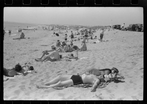 A black and white photo of a busy beach with people sunbathing, relaxing, and enjoying the sand and sea.