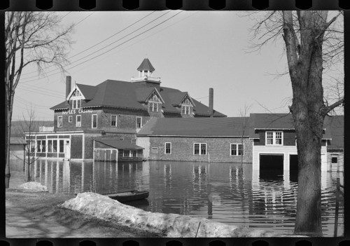 Historic black-and-white photo of a building partially submerged in floodwaters, with trees and power lines visible.