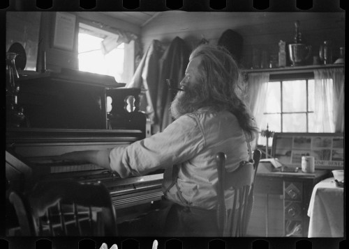 A man with long hair and a beard sits at a piano in a cozy, cluttered room with sunlight streaming in.