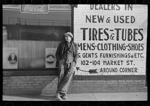 A man in worn clothing leans against a wall with a sign advertising tires, clothing, and sandwiches.