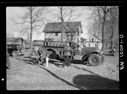 A vintage fire truck parked near a house, with firefighters connecting hoses to a hydrant on a grassy area.