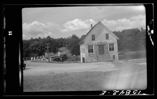 A vintage black-and-white photo of a small building by a river, with a dock and trees in the background.