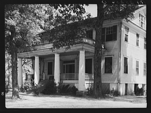 Historic two-story house with a large porch, supported by columns, surrounded by trees and greenery.