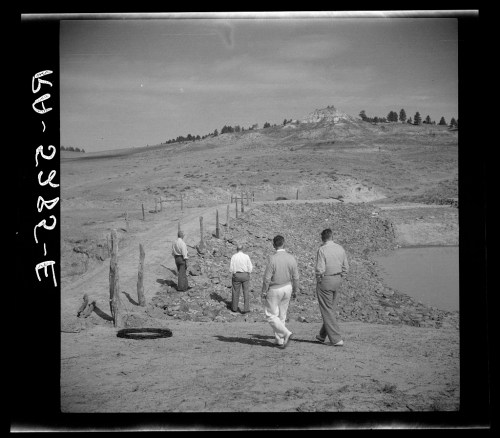 Four men walking along a dirt path near a small pond, with a hilly landscape in the background.