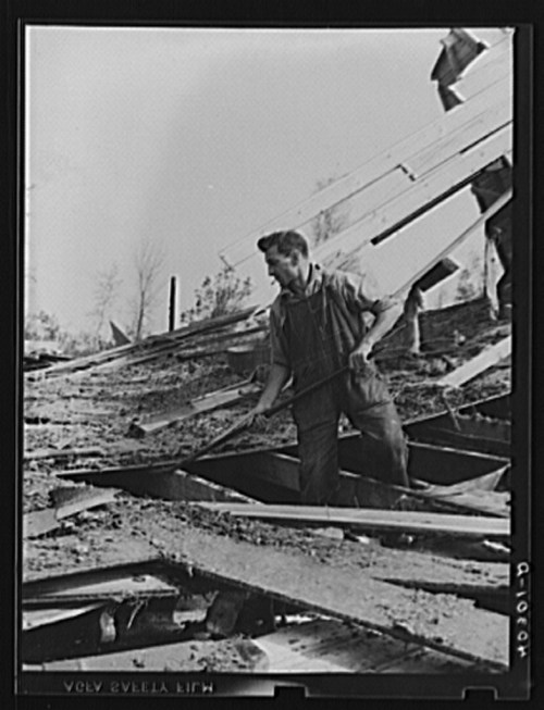 A man shovels dirt amidst debris from a collapsed structure, surrounded by trees and a clear sky.