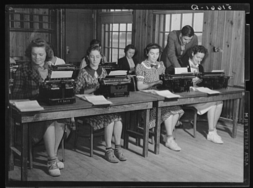 A group of women typing on typewriters in a classroom setting, with a man assisting in the background.