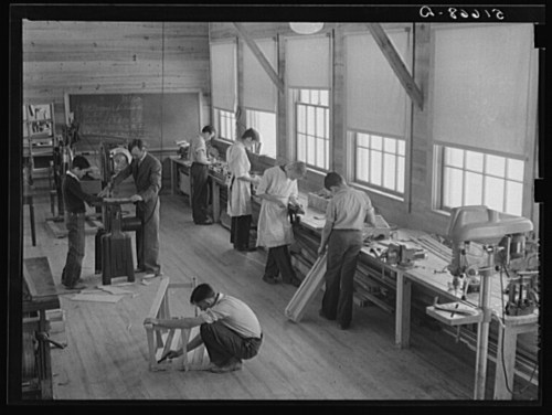 A workshop scene with several men working on various woodworking projects in a well-lit space.