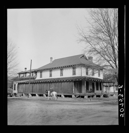 Historic two-story house with a porch, surrounded by trees and a dirt road, featuring a horse nearby.