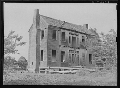 Abandoned two-story house with boarded windows and a deteriorating facade, surrounded by trees and a wooden fence.