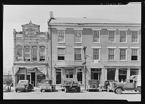 Historic storefronts with vintage cars parked outside, featuring a grocery store and a two-story building.