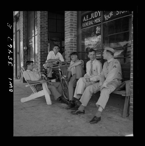 Five young men sit outside a general store, chatting and relaxing on a sunny day, with a bicycle nearby.