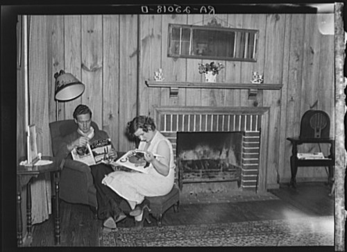 A man and woman sit in a cozy room, reading magazines by a fireplace with wooden walls and a decorative mirror.