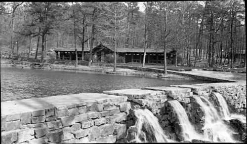 A serene black-and-white scene of a stone wall with waterfalls, surrounded by trees and a rustic building by a lake.
