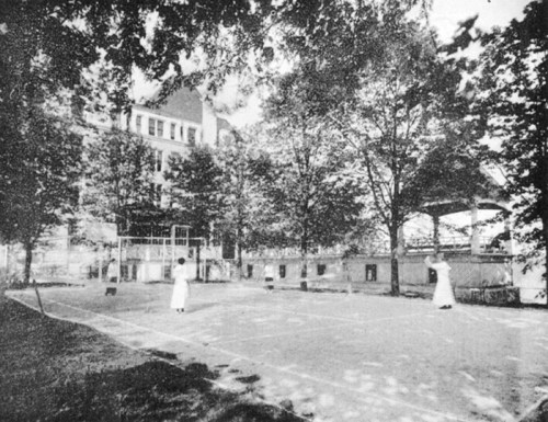 Two women in white dresses play tennis on a court surrounded by trees and a building in the background.