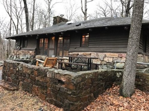 A rustic cabin made of stone and wood, surrounded by trees and fallen leaves, with a small porch and outdoor seating.