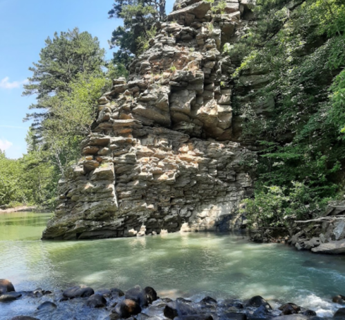 A rocky cliff beside a calm river, surrounded by lush green trees under a clear blue sky.