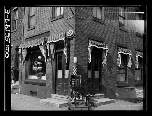 Black and white photo of a corner storefront with decorative awnings and a barber pole outside.