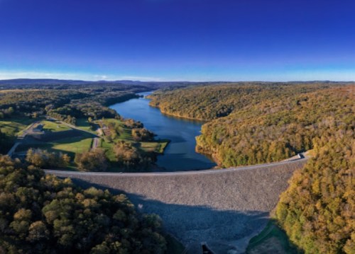 Aerial view of a dam surrounded by lush green hills and a serene lake under a clear blue sky.