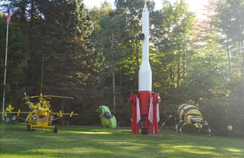 A whimsical outdoor scene featuring a yellow helicopter, a rocket, and large insect sculptures in a grassy area.